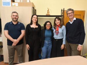 Five people pose for a photograph in a room at the University of Ferrara. From left to right are Professor Andrea Rubin, master's student Giovanna Maria de Souza Almeida, master's student Paula Kaliane Henrique Silva, Professor Maria Giovanna Guedes Farias, and Professor Marco Bresadola. The image was taken during the welcoming meeting for the PPGCI/UFC master's students participating in an academic mobility program at the Italian institution.