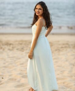 Foto em plano aberto de Andreza em uma praia. Ela usa um vestido longo de alcinhas na cor azul-claro, com cabelos soltos e um sorriso amplo, em uma pose descontraída de costas com o rosto virado para a câmera.